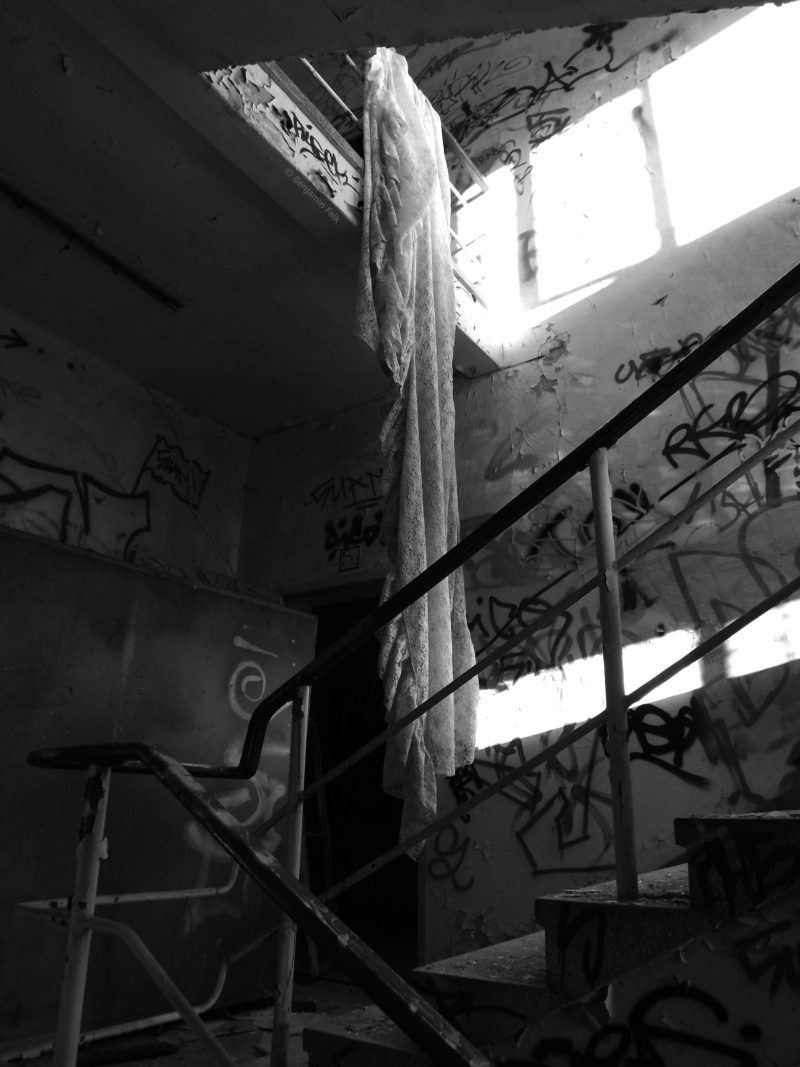 Schwarz-Weiß-Fotografie eines abbruchreifen und mit Graffiti beschmierten Treppenhauses, in dem eine Gardine von oben herunter hängt.