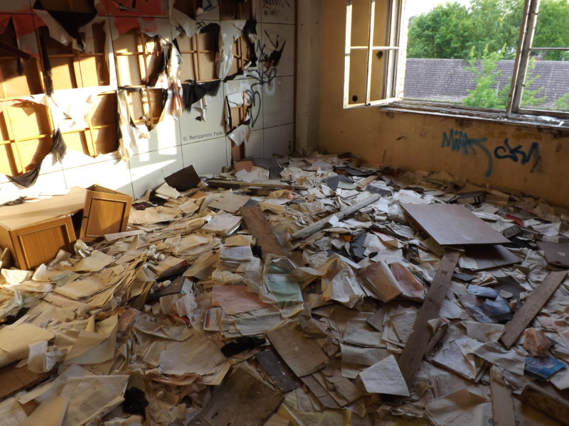 Raum mit zerrissenen Wandbehängen, dessen Boden mit kaputten Akten übersäht ist.