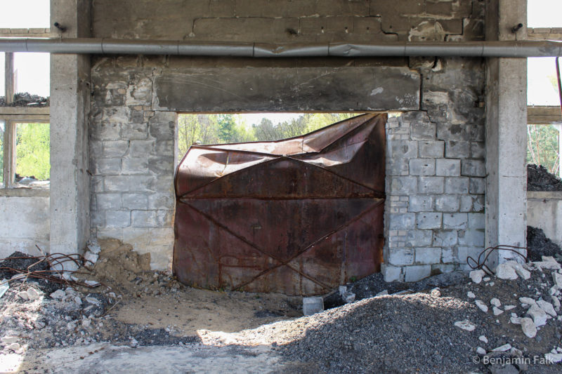 Verbogenes und verrostetes Metalltor in einer eingedrückten Beton-Stein-Wand hinter herumliegendem Schutt.