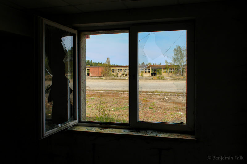Blick aus einem dunklen Raum heraus, durch ein Fenster mit zerbrochenen Scheiben, auf einen verlassenen Werkshof mit dahinterstehender Halle.