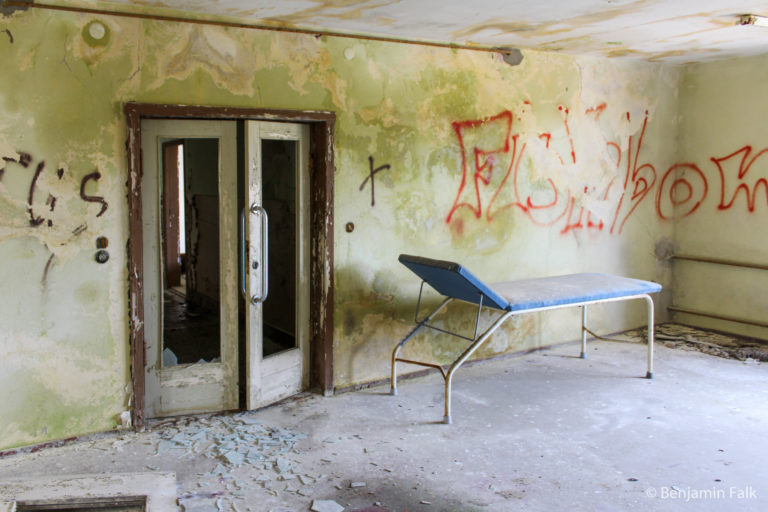 Leerstehender Raum mit einer doppelten FLügeltür, mit herausgebrochenem Glas und einer staubigen Krankenliege, vor einer Wand mit Grünspan undfi.