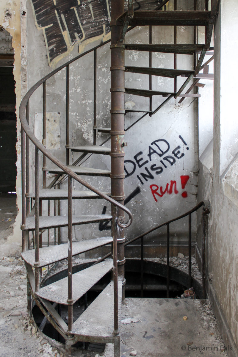 Wendeltreppe mit Schutt und Staub bedeckt, die von oben nach unten führt. Hinter der Treppe ist ein Graffitti "DEAD INSIDE! Run!" mit einem Pfeil nach unten.
