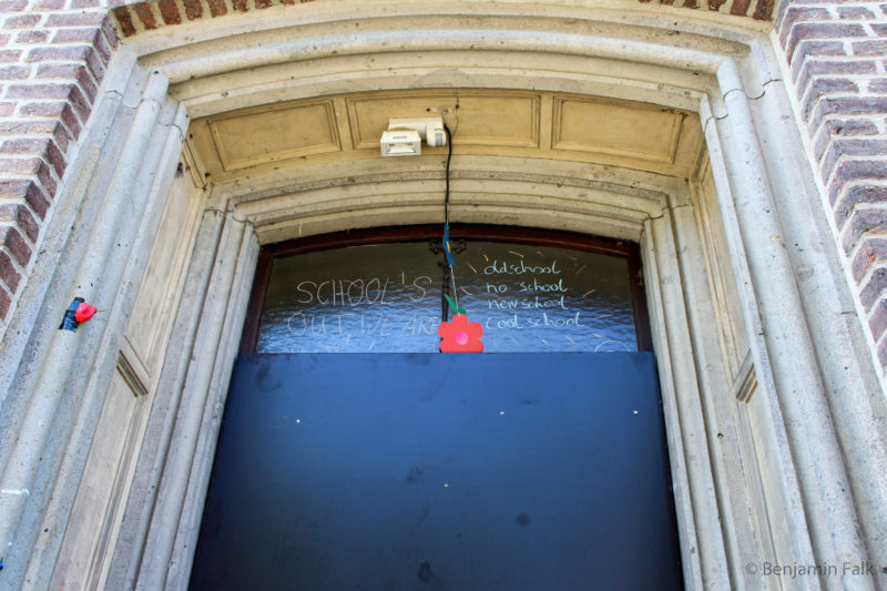 Mit Brettern verschlossene Tür eines Backsteinbaus, über dem Brett ein Fenster auf dem steht "SCHOOLS OUT" und old school, no school, new school, cool school"