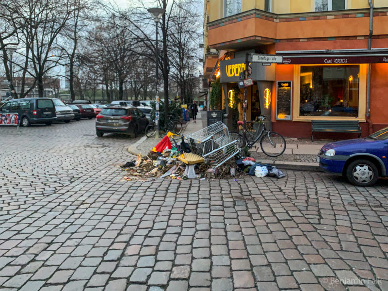 Ein Sperrmüllhaufen mit einem Einkaufswagen auf einer Straßenkreuzung an der Berliner Sonntagsstraße, vor einem Cafe.