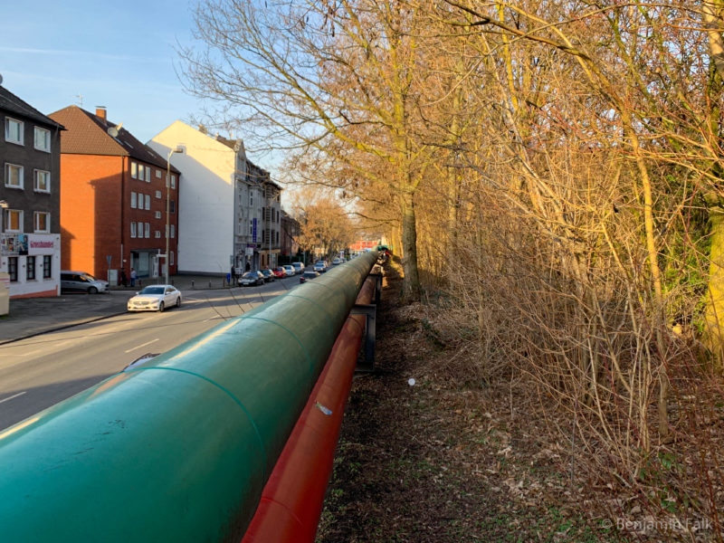 Fernwärme-Rohre auf der einen Seite an einer Hauptstraße, auf der anderen Seite an einem Gebüsch vorbei verlaufend.