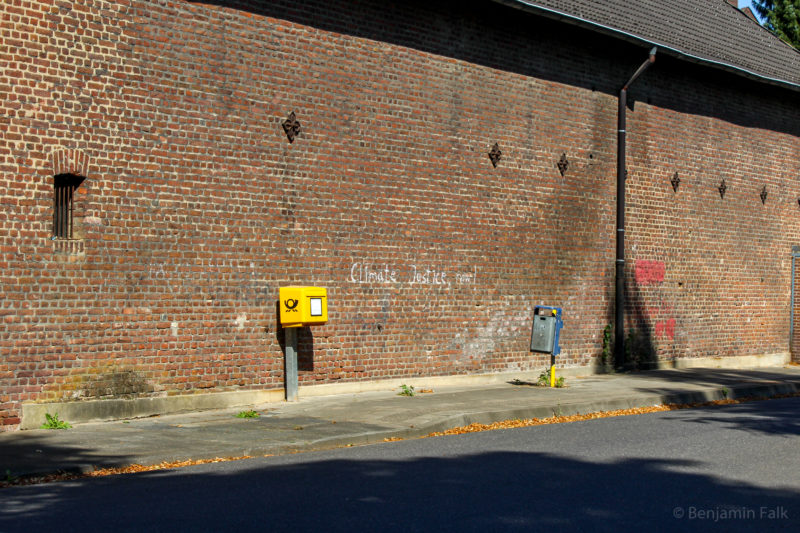 Schiefstehender Briefkasten auf einem zugewachsenen Bürgersteig vor einer Backstein-Mauer mit dem Graffiti "Climate Justice Now"