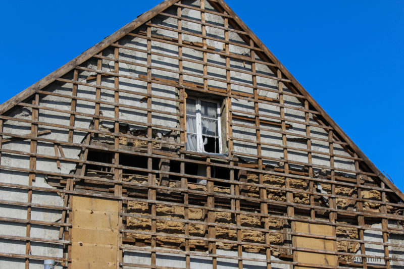 Entkleidete Fassade mit freiliegendem Holzgerippe vor blauem Himmel und einem einzelnen Fenster mit einer weißen Gardine.
