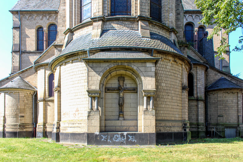 Kruzifix an der Außenwand der Rückseite einer katholischen Kirche, mit Graffiti darunter, mit der halben Kirche im Bildausschnitt.