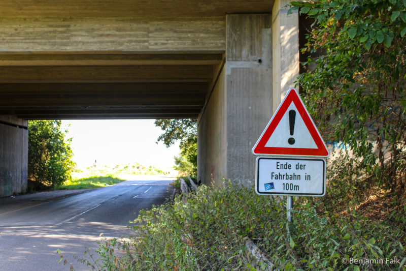 Straßenschild "Ende der Fahrbahn in 100m" am zugewachsenen Rand einer Straße vor einer Autobahn-Brücke.