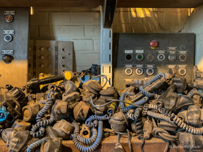 Ein Haufen abgetrennter staubiger und dreckiger Telefonhöhrer in einem Regal, vor einer Reihe von demontierten Schaltbrettern.