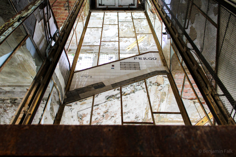Das innere eines ehemaligen verglasten Afzugschachtes in der Mitte eines Treppenhauses, mit zerbrochenenen Scheiben und ablätternden Fliesen und Wandfarben, mit Graffiti und rostigen Stahlträgern.