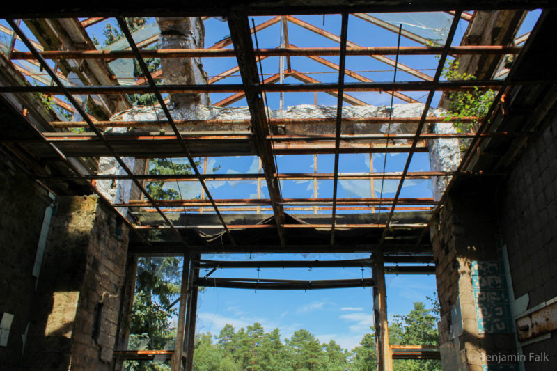 Glasdach mit doppeltem Spitzdach, bei denen das Glas zerbrochen ist und die Stahlträger und Lüftungsschächte offen vor einem blauen Himmel liegen.