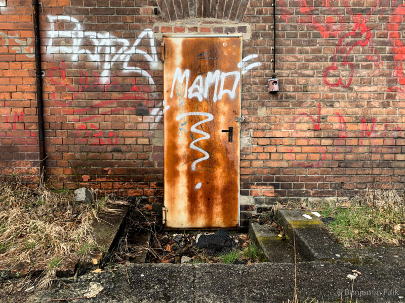Rostige Eisentüür in einer Bachsteinwand eines Bahn-Betriebs-Gebäudes, mit Graffiti und elektrischen Leitungen daneben