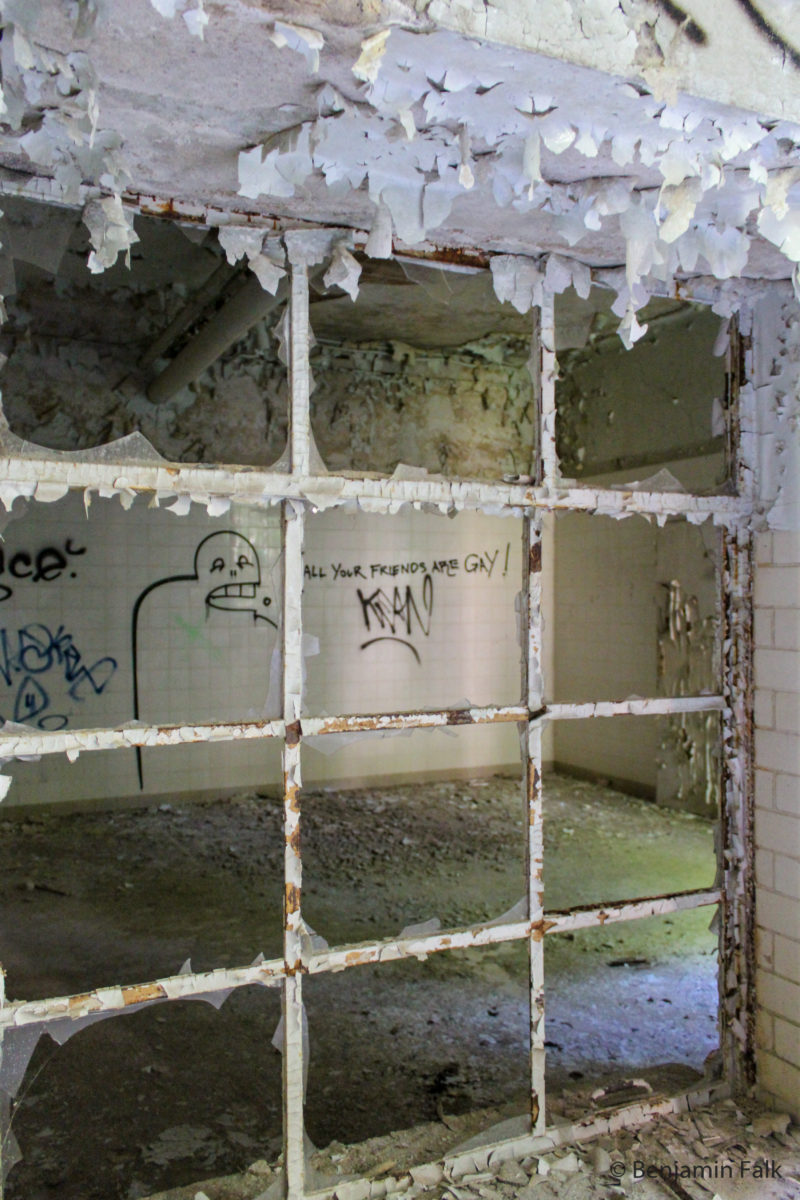 Blick durch den Fensterrahmen eines entglasten Fensters, in einen weiß gefliesten Raum mit Grafitti.