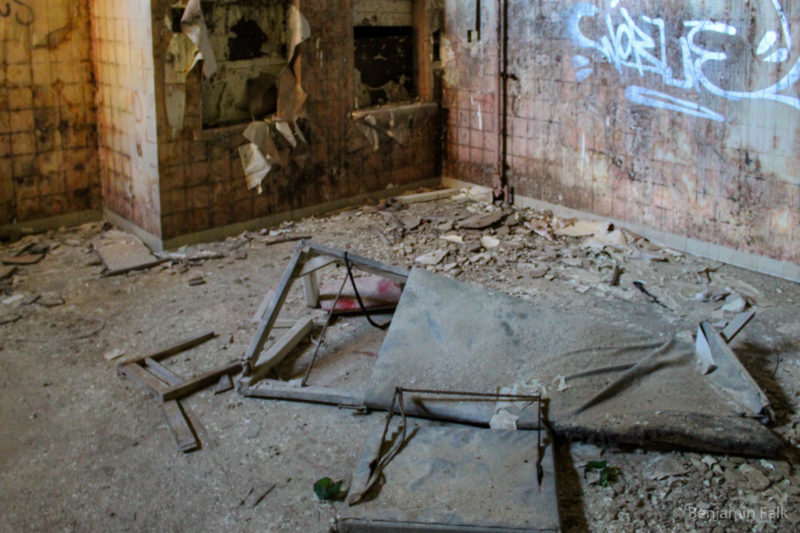 Polsterfläche einer medizinischen Liege in Einzelteilen auf einem schutt-übersähtem Boden vor einer braun gefliesten dreckigen Wand mit Graffiti.