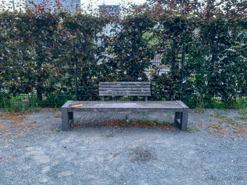 Leere Holz-Parkbank vor einem Zaun mit einer Hecke auf der ein einzelnes Buch liegt.