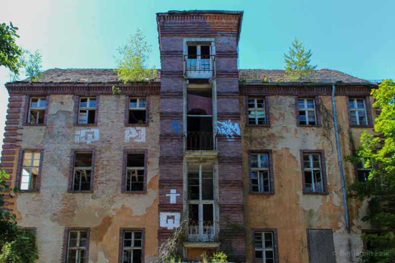 Mehrstöckiges verlassenes Backstein-Gebäude, frontal vor einem blauem Himmel aufgenommen. Zwischen den Etagen sind vereinzelt Graffitis, die Fenster sind größtenteils entglast und teilweise mit Brettern vernagelt.