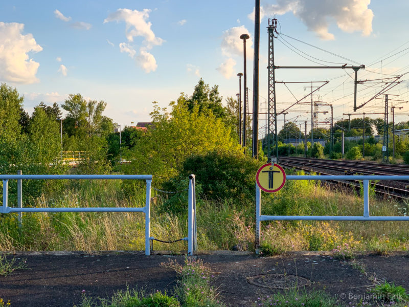 Ende eines zugewachsenen Bahnsteigs mit einer Absperrung und einem "Durchgang verboten" Symbol, mit Blick auf einen verwilderten Grünstreifen in einem Eisenbahn-Betriebsgelände mit Stromleitungen und Laternen
