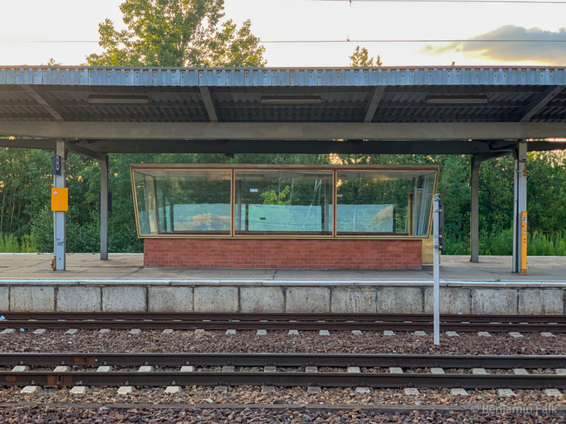 Blick über die Bahngleise hinweg auf einen Bahnsteig mit einem leeren Wartehäuschen, mit einem backsteinfundament und Verglasung unter einem Wellblechdach.