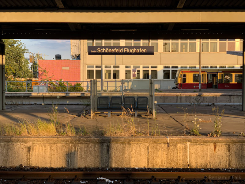 Bahnsteig auf dem Gras wächst, mit Blick auf 4 Stahlsitze in einem Winschutz mitten auf dem Bahnsteig, während auf dem zweiten Bahnsteig dahinter eine S-Bahn mit offenen Türen wartet. Fotografiert im Licht des Sonnenuntergangs.