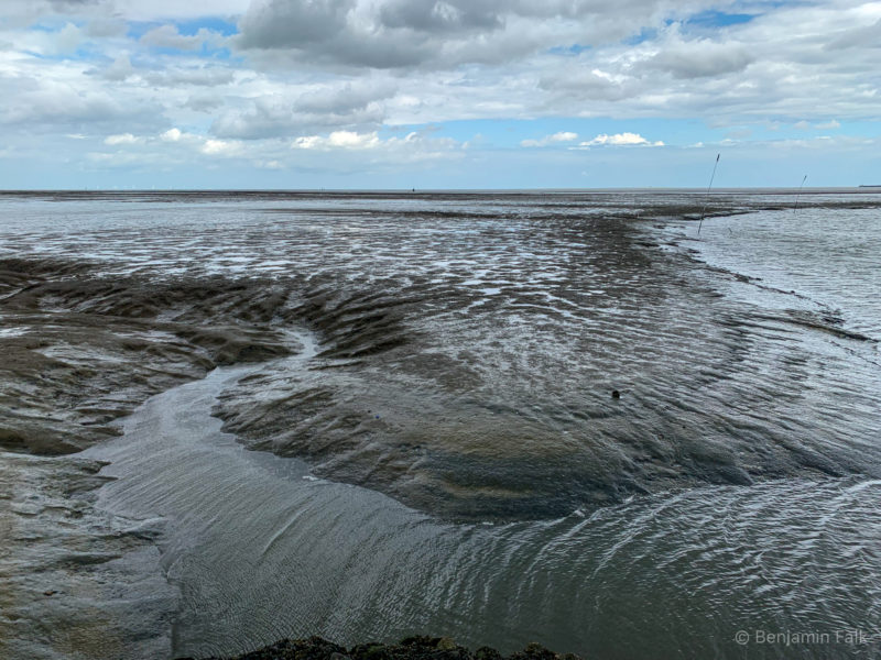Abfließendes Wasser im Wattenmeer, vor einem leicht bewölktem blauem Himmel. Das Wasser bildet Muster auf dem Boden.