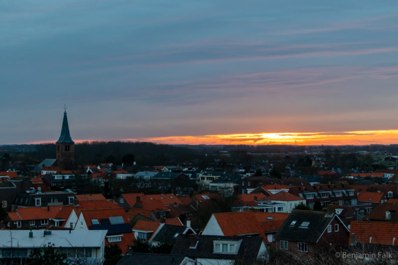 Blick über die roten Dächer des Bad Domburgs mit dem Kirchturm im Licht der roten Morgensonne, die durch ein Wolkenband scheint.