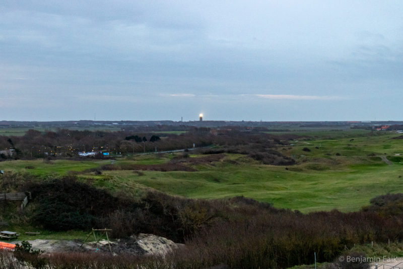 Dünenlandschaft über einen Spielplatz und eine Siedlung zwischen grünen Hügeln hinweg mit einen leuchtendem Leuchtturm am Horizont.