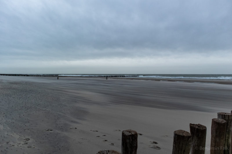 Strand bei Ebbe mit einer aufgewühlten See am Horizont, im Sand die Spuren von Wind und einzelne Fußabdrücke.