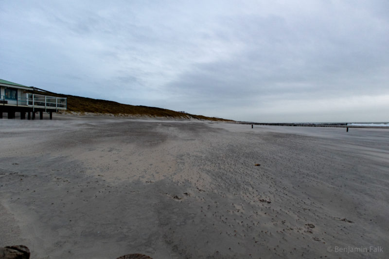 Strand mit Muscheln über die der Wind hinweg geblasen hat. Links Dünen, rechts Wellenbrecher und Wellen. Der Himmel ist bewölkt.