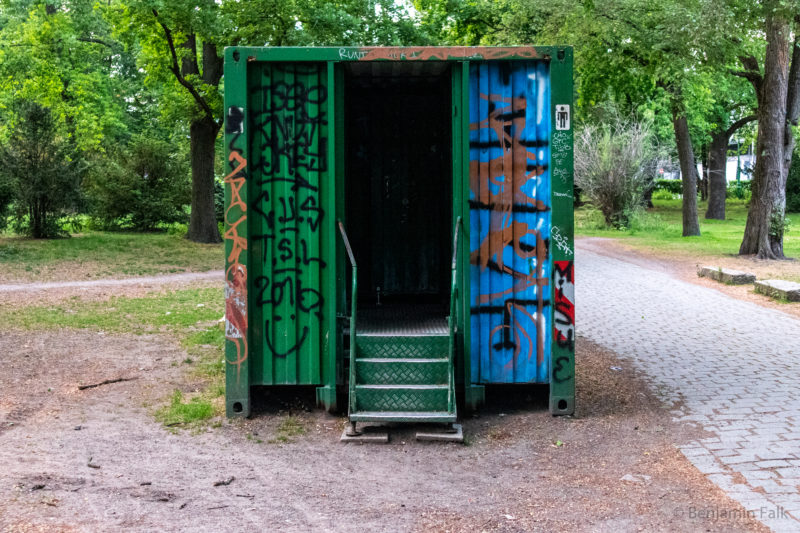 Öffentlicher Toilettencontainer im Tiergarten, beschmiert mit Graffiti, Blick frontal auf den Eingang, der komplett dunkel ist.