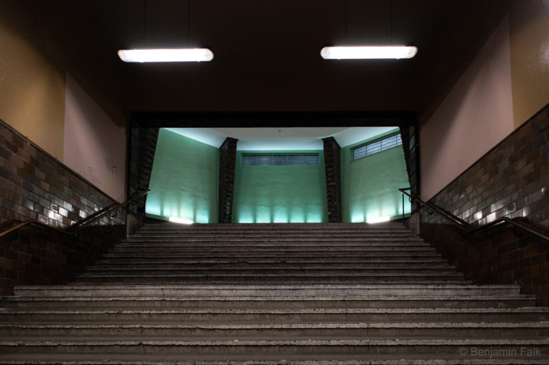 Braun gefliester Treppenaufgang des S Wannsee mit Blick in die beleuchtete Bahnhofshalle mit den Grünen Wänden.
