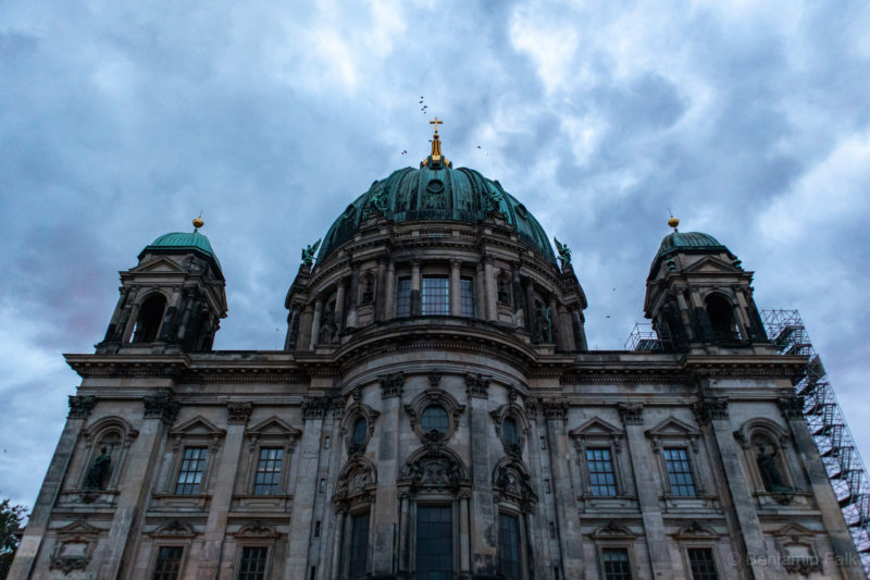 Rückseite des Berliner Doms, nach oben blickend aufgenommen vor einem bewölkten Himmel mit darüber fliegenden Krähen