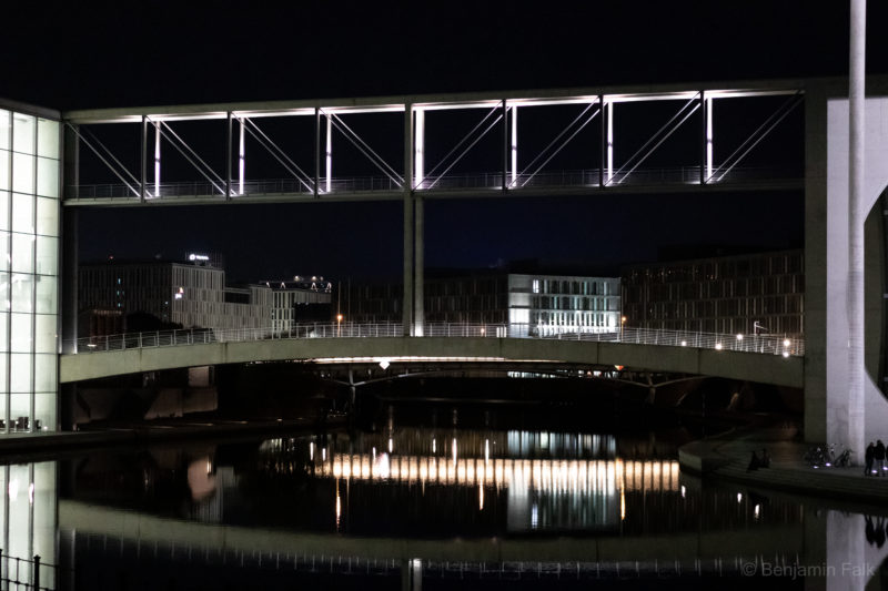 Beleuchtete Spreebrücke zwischen den Bundestagsgebäuden bei Nacht mit Reflexion über der Spree, aufgenommen von Höhe des Reichstagsgebäudes aus.