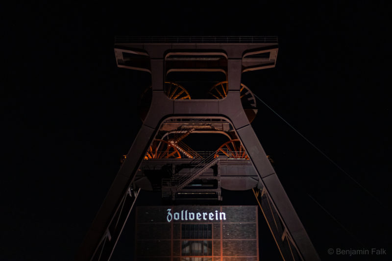 Beleuchteter Turm der Zeche Zollverein bei Nacht, fotografiert frontal von unten mit deutlich herausleuchtendem "Zollverein"-Schriftzug.