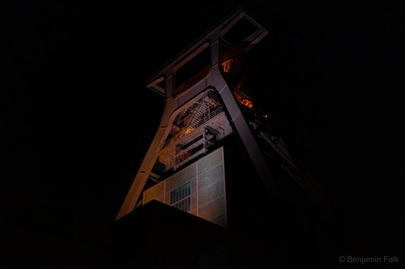 Rückseite des beleuchteten Turms der Zeche Zollverein bei Nacht vor komplett schwarzem Hintergrund.