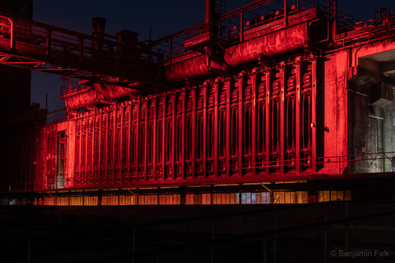 Verlassenes Industriegebäude mit verschiedenen Versorgungsrohren und einem Stahlgerippe n der Seite, aufgenommen bei Nacht mit komplett rot angestrahltem Stahlgerippe.