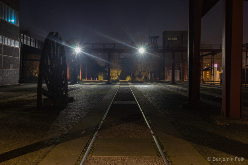 Schienenstrang auf einem angestrahlten verlassenen platz bei Nacht, der zwischen Strahlträgern und einem Schwungrad eines Förderturms hinweg auf den Platz verläuft.