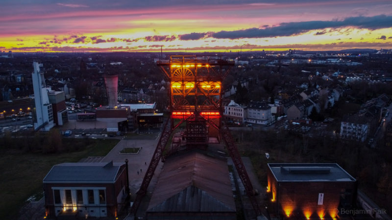 Drohnenfoto des roten Stahl-Förderturms der Zeche Consolidation (Gelsenkirchen) in der Dämmerung, bei beleuchtetem Förderturm und Gebäuden, mit dem Sonnenuntergang über dem Stadtpanorama im Hintergrund.