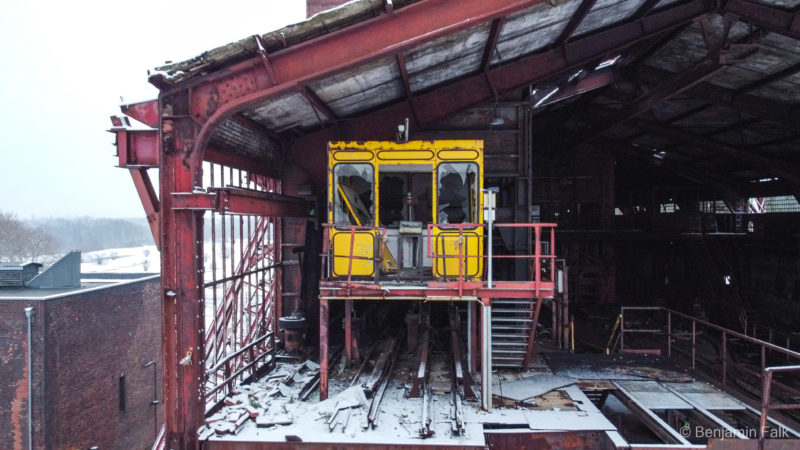 Blick auf eine seitlich offene Industriehalle aus rotem Stahl, mit Fokus auf eine gelbes Schalthäuschen, auf Säulen über zwei Bahnschienen.