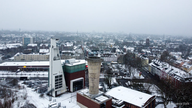 Luftaufnahme des verschneiten Ruhrgebiets. Im Vordergrund ist der Kühlturm des Consol-Theaters zu sehen, dahinter ein Personen-Förderturm. Der Blick führt über die Stadt mit Nebel am Horizont und verschneiten Dächern.
