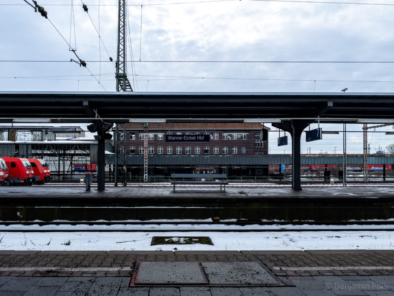 Foto des zugeschneiten Bahnhofs Wanne-Eickel Hbf, mit Blicka uf eine verlassene Bahnhofsbank, über die zugeschneiten Schienen hinweg. Hinter der Bank sind verschiedene Bahngebäude, sowie 4 wartende rote Loks nebeneinander zu sehen.