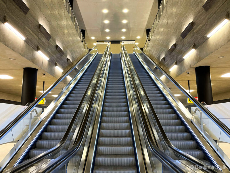 Rolltreppen des U-Bahnhofs unter den Linden, fotografiert mit Blick auf der mittleren Rolltreppe von unten nach oben, mit den Rolltreppen die durch die Steinverkleidete Decke auf die Lichter der Verteilerebene zu führen.
