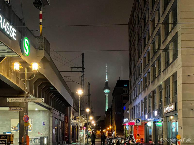 Straßenfoto vom Bahnhof Friedrichstraße in Berlin, aufgenommen die Parralelstraße an der Bahnbrücke entlang mit Blick auf den Fernsehturm. Das Bild ist nachts, mit leuchtender Straßenbeleuchtung aufgenommen, bei geschloseneen Geschäften. Der Fernsehturm hebt sich beleuchtet vom Bewölkten Nachthimmel ab.