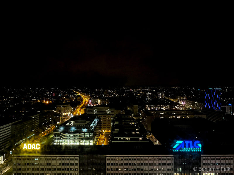Foto vom Stadtpanorama aus einem Hochhaus aufgenommen, vom Berliner Alexanderplatz weg mit Blick auf den ADAC bei Nacht. Im Vordergrund ist das Hochhaus zu erkennen, während daneben die Lichter der Autos auf der Straße nach hinten verschwinden und kleiner werden. Von den Häusern der Stadt sind nur die beleuchteten Fenster und Straßen zu erkennen.
