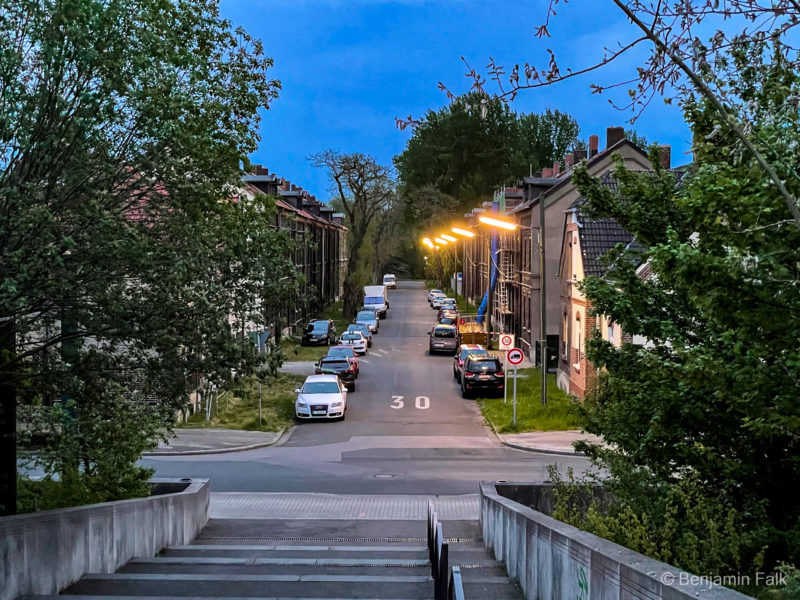 Straße mit alten Bergarbeiterhäusern in der blauen Stunde, beleuchtet von Natrium-Lampen. Fotografiert vom oberen Ende einer Treppe, die vollgeparkte Straße, mit zuweachsenenen Gehwegen entlang mit Blick auf eine Wand aus Bäumen am Ende.