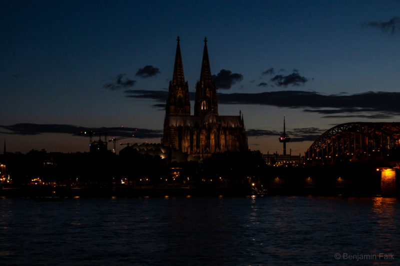 Aufnahme nach Sonnenuntergang über den Rhein hinweg mit Blick auf den Kölner Dom. Die Lichter des Doms und der Hohenzollern-Brücke leuchten über den Rhein hinweg, vor einem leicht bewölkten Himmel mit dem Restlicht der untergegangenen Sonne. Abgesehen von den Lichtern sind die Wolken und Gebäude nur noch als schwarze Schatten zu erkennen.