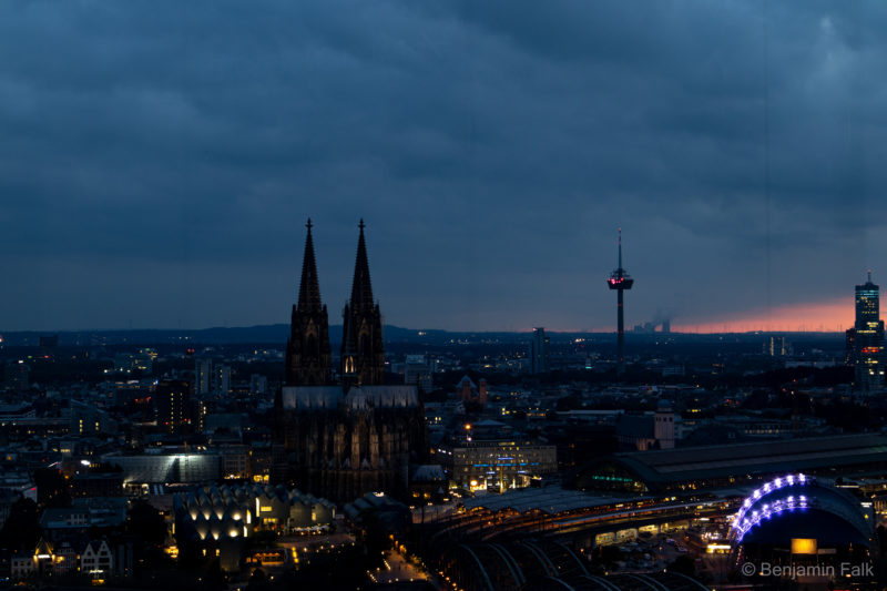 Aufnahme von der gegenüberliegenden Rheinseite vom Aussichtsturm auf Dom und Bahnhof gerichtet, über die Stadt hinweg mit eingeschalteteer Beleuchtung der Innenstadt und des Bahnhofs im Licht der Dämmerung.