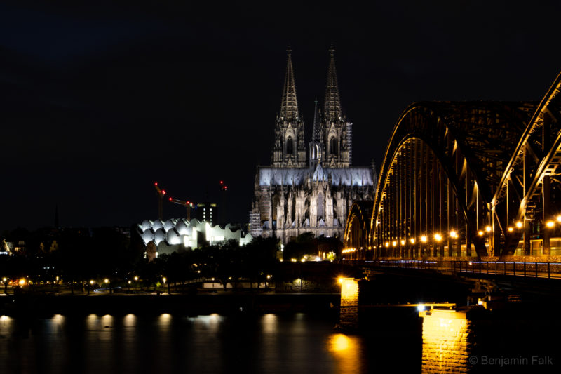 Aufnahme vom Kennedy-Ufer entlang der Hohenzollern-Brücke gerichtet auf den angestrahlten Kölnder Dom Kölner Dom und die Philharmonie bei Nacht in einer Langzeitbelichtung mit der Bewegungsunschärfe des Rheins und der über die Brücke fahrenden Züge.