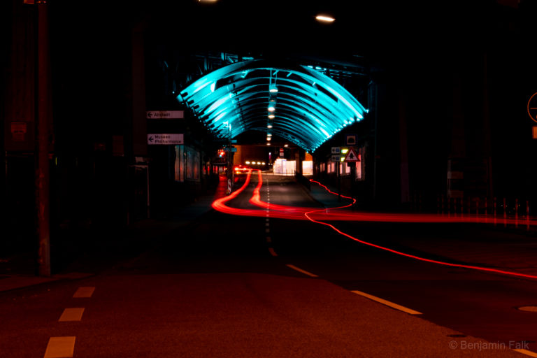 Langzeitbelichtung eines Straßentunnels unter einer Eisenbahnbrücke (Trankgasse, Köln). Die Stahlträgerkonsturktion an der Decke des "Tunnels" ist blau angestrahlt, auf der Straße zeichnen sich die Lichtspuren von Autos und Fahrrädern ab.