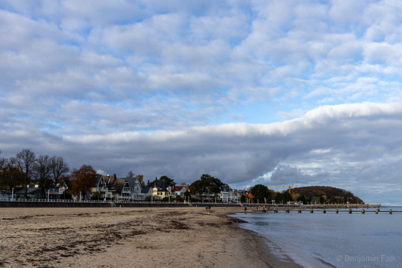 Travemünder Strand, forografiert von der Wasserkante entland des Wassers auf die Strandpromenade. Über den Bäumen und Häusern der Promenade liegt ein Wolkenwand vor einem blauen Himmel.
